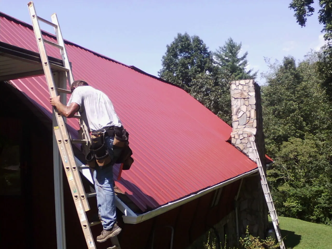 Roofer on a ladder beside a red metal roof and stone chimney — Soffit & Fascia Repair by Roofing Brothers in Tehama